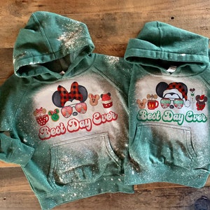 May include: Two green hooded sweatshirts with a bleach-splattered design. The sweatshirts feature a Christmas-themed graphic with Mickey Mouse and friends, along with the text "Best Day Ever".