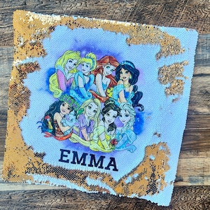 May include: A white and gold sequin pillow with a purple background featuring a group of Disney princesses including Cinderella, Snow White, Belle, and Ariel. The pillow has the name "EMMA" printed in black letters.