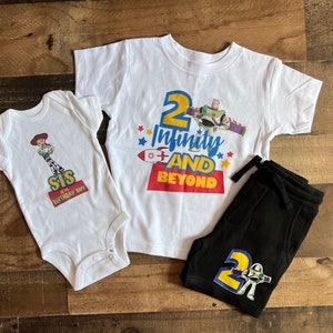 May include: Two white t-shirts with a Toy Story theme. One shirt says "2 Infinity and Beyond" with a picture of Buzz Lightyear. The other shirt says "Sis is the Birthday Boy" with a picture of Jessie. A pair of black trousers with a Buzz Lightyear graphic and the number 2.