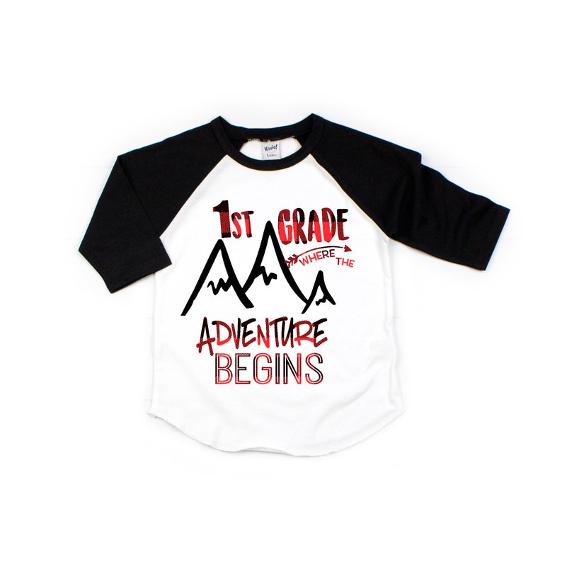 May include: White and black baseball tee shirt with red and black mountain graphic. The text on the shirt reads "1st Grade Where the Adventure Begins".