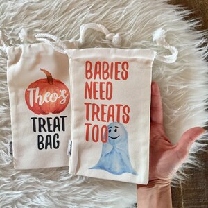 May include: Two white canvas treat bags with orange and black text. One bag has a pumpkin with the text "Theo's Treat Bag". The other bag has a blue ghost with the text "Babies Need Treats Too".