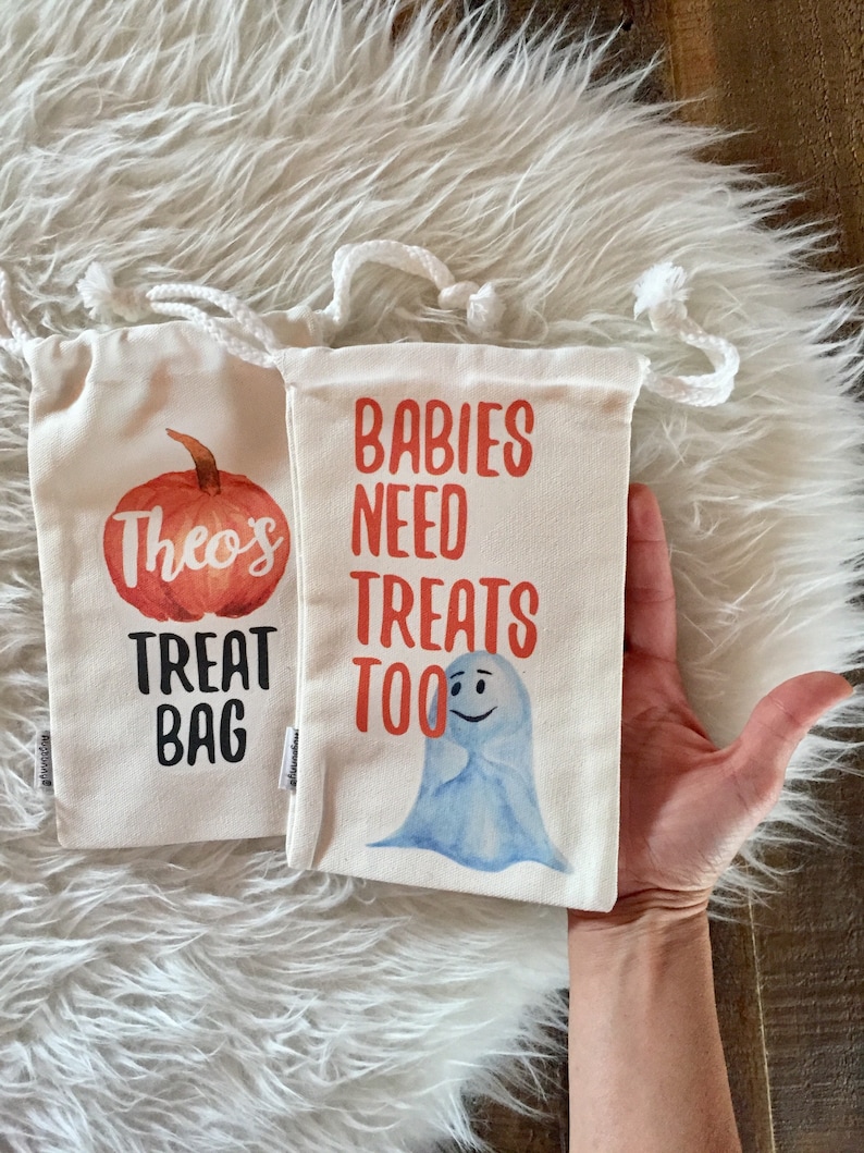 May include: Two white canvas treat bags with orange and black text. One bag has a pumpkin with the text "Theo's Treat Bag". The other bag has a blue ghost with the text "Babies Need Treats Too".