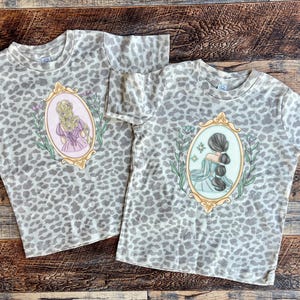 Peut inclure: Deux t-shirts &agrave; imprim&eacute; l&eacute;opard gris et blanc avec une illustration vintage d'une femme aux cheveux longs dans un cadre. L'illustration sur le t-shirt de gauche repr&eacute;sente une femme aux cheveux blonds et une robe rose. L'illustration sur le t-shirt de droite repr&eacute;sente une femme aux cheveux bruns et une robe bleue.