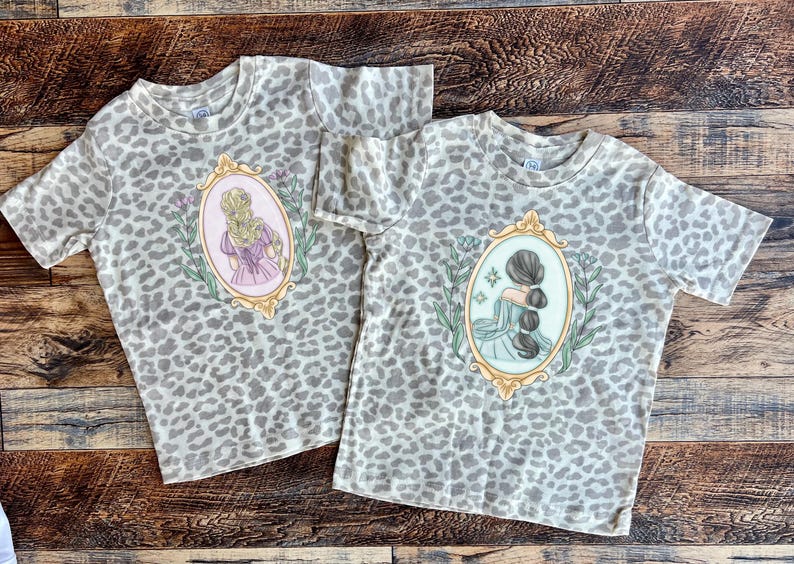 Peut inclure: Deux t-shirts &agrave; imprim&eacute; l&eacute;opard gris et blanc avec une illustration vintage d'une femme aux cheveux longs dans un cadre. L'illustration sur le t-shirt de gauche repr&eacute;sente une femme aux cheveux blonds et une robe rose. L'illustration sur le t-shirt de droite repr&eacute;sente une femme aux cheveux bruns et une robe bleue.