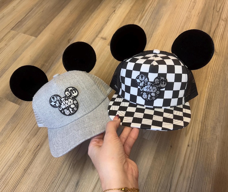 May include: Two Star Wars themed baseball caps with black Mickey Mouse ears. One cap is grey with a Star Wars logo, the other is black and white chequered. Both caps have a mesh back.