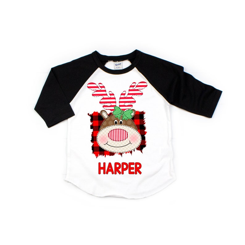 May include: White and black baseball tee shirt with a red and black plaid background and a cartoon reindeer with a green bow. The text "HARPER" is printed below the reindeer.