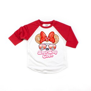 May include: White and red baseball tee shirt with a cartoon image of Minnie Mouse wearing sunglasses with a reflection of Cinderella's castle. The text "Best Day Ever" is printed in pink below the image.
