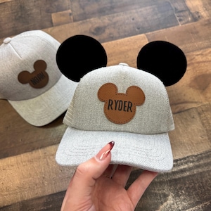 May include: Two grey baseball caps with brown Mickey Mouse ear patches. One patch says "Noah" and the other says "Ryder". The cap in the foreground has large black ears attached to the top.
