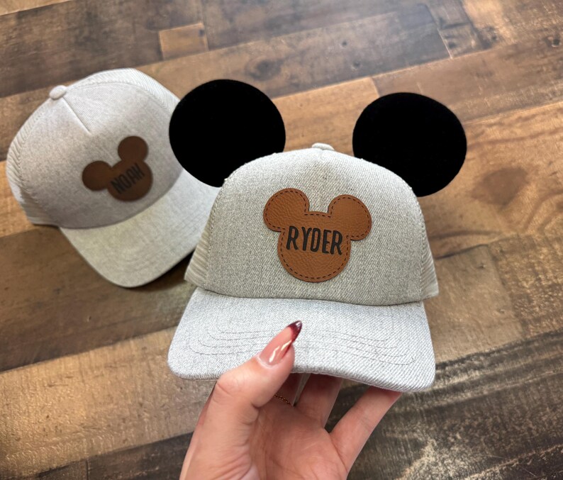 May include: Two grey baseball caps with brown Mickey Mouse ear patches. One patch says "Noah" and the other says "Ryder". The cap in the foreground has large black ears attached to the top.