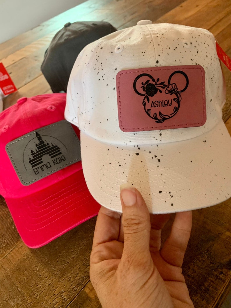 Pu&ograve; includere: Due cappellini da baseball personalizzati. Uno &egrave; rosa con una grafica di un castello bianco e il testo "Emma Kate". L'altro &egrave; bianco con macchie nere e una toppa rosa con una silhouette di Topolino nera e il testo "Ashley".