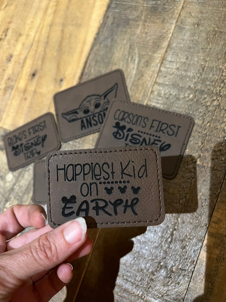 Pu&ograve; includere: Quattro toppe in pelle marrone con testo nero. Le toppe dicono "Riko's First Disney Trip", "Anson", "Corson's First Disney", e "Happiest Kid on Earth".