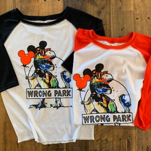 May include: Two white t-shirts with black and red sleeves. The shirts feature a colorful graphic of a dinosaur wearing Mickey Mouse ears and sunglasses, holding a red balloon. The text "WRONG PARK" is printed on the shirts.