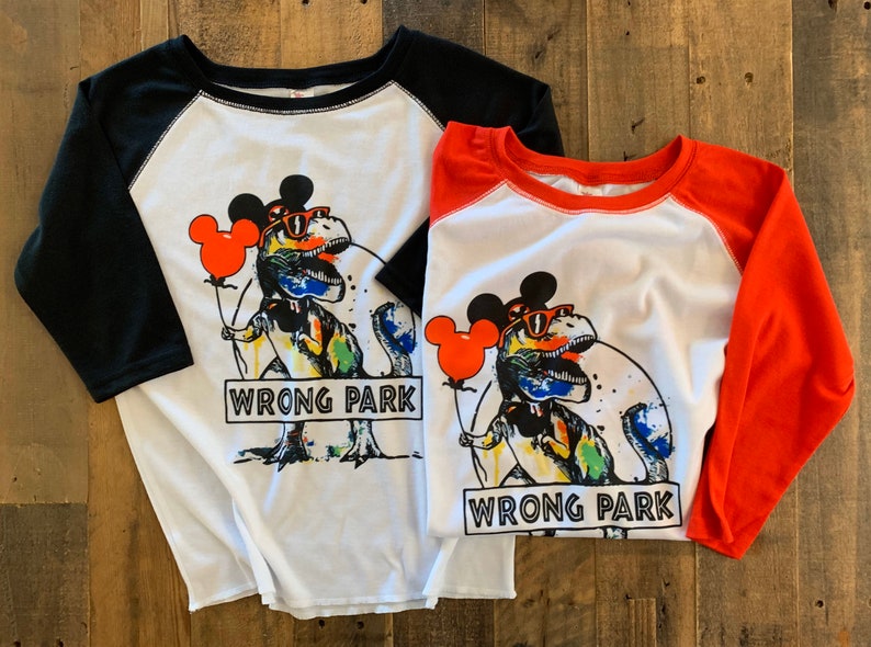 May include: Two white t-shirts with black and red sleeves. The shirts feature a colorful graphic of a dinosaur wearing Mickey Mouse ears and sunglasses, holding a red balloon. The text "WRONG PARK" is printed on the shirts.