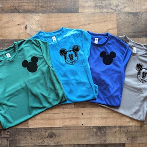 May include: Four short-sleeved t-shirts in shades of teal, blue, and gray, each featuring a Mickey Mouse graphic. Two shirts have a solid black Mickey Mouse head, while the other two have a sketched design. The shirts are laid out on a wooden surface.