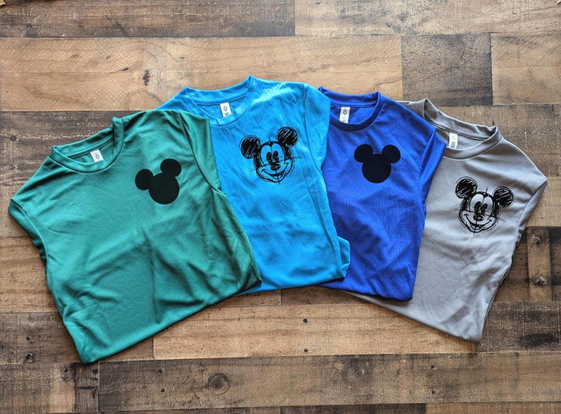 May include: Four short-sleeved t-shirts in shades of teal, blue, and gray, each featuring a Mickey Mouse graphic. Two shirts have a solid black Mickey Mouse head, while the other two have a sketched design. The shirts are laid out on a wooden surface.