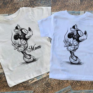 May include: Two white t-shirts with a black and white illustration of Mickey Mouse and Minnie Mouse. The shirt on the left says "Mom" on the bottom right corner.
