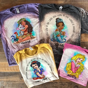 Peut inclure: Quatre t-shirts blanchies avec des motifs de princesses Disney. Les t-shirts sont violet, noir, jaune et rose. Les motifs incluent Moana, Tiana, Blanche-Neige et Aurore. Les t-shirts ont des &eacute;claboussures de javel et les noms des princesses sont &eacute;crits sous les motifs.
