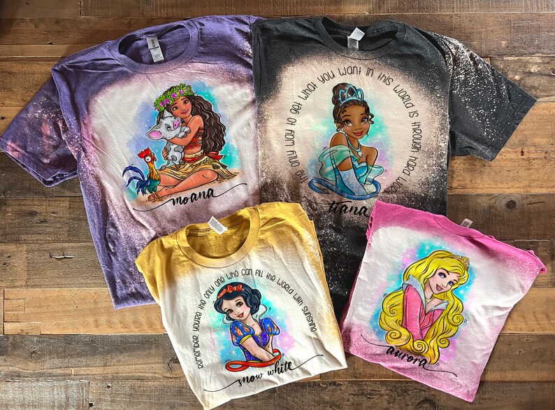 Peut inclure: Quatre t-shirts blanchies avec des motifs de princesses Disney. Les t-shirts sont violet, noir, jaune et rose. Les motifs incluent Moana, Tiana, Blanche-Neige et Aurore. Les t-shirts ont des &eacute;claboussures de javel et les noms des princesses sont &eacute;crits sous les motifs.