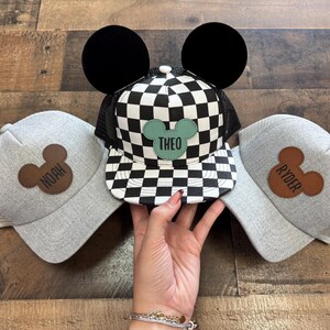 May include: Three baseball caps with Mickey Mouse ear designs. One cap has a black and white chequered pattern with the name "THEO" on a green patch. The other two are grey with brown Mickey Mouse patches that say "NOAH" and "RYDER".