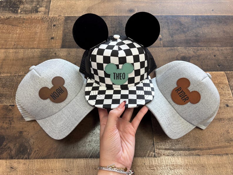 May include: Three baseball caps with Mickey Mouse ear designs. One cap has a black and white chequered pattern with the name "THEO" on a green patch. The other two are grey with brown Mickey Mouse patches that say "NOAH" and "RYDER".