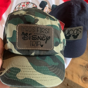 May include: A camouflage baseball cap with a brown leather patch reading "THEO'S FIRST DISNEY TRIP". A navy blue cap with "lucas" and Mickey Mouse silhouettes is also visible, along with a red and white raglan shirt.