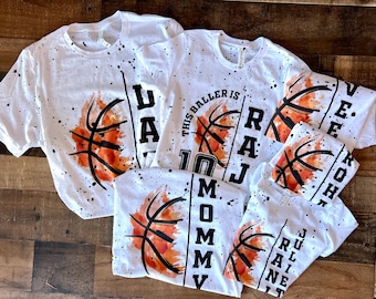 May include: A collection of white t-shirts featuring a black paint splatter pattern, a watercolor-style orange basketball graphic, and black text. The shirts include personalized names such as Raj, Mommy, and Julian, with the text This Baller Is.