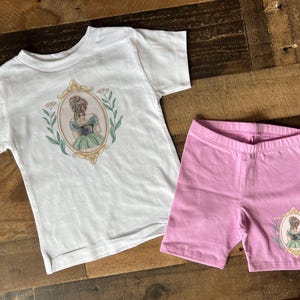 Peut inclure: Un t-shirt blanc avec un motif floral et une fille en robe verte dans un cadre. Un short rose avec un motif floral similaire et une fille en robe verte dans un cadre.