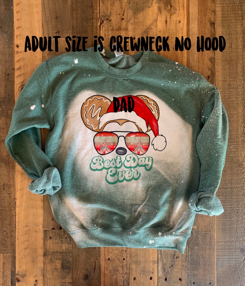 May include: Green sweatshirt with a bleached effect featuring a cartoon Mickey Mouse head with a Santa hat and sunglasses. The sunglasses reflect the Cinderella Castle. The text "Dad" is on the hat and "Best Day Ever" is below the sunglasses.