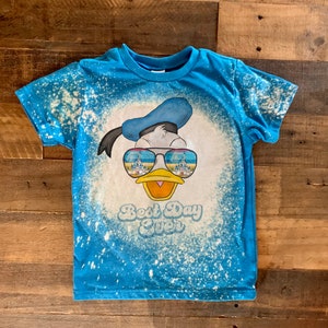 Puede incluir: Una camiseta azul con un dise&ntilde;o salpicado de lej&iacute;a que presenta un dibujo animado de Donald Duck con gafas de sol con un reflejo de castillo. El texto "Best Day Ever" est&aacute; impreso debajo de la imagen.