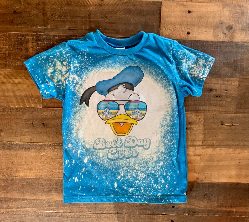Puede incluir: Una camiseta azul con un dise&ntilde;o salpicado de lej&iacute;a que presenta un dibujo animado de Donald Duck con gafas de sol con un reflejo de castillo. El texto "Best Day Ever" est&aacute; impreso debajo de la imagen.