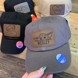 May include: Four baseball caps with personalized patches. The caps are black, beige, gray, and black. The patches feature a cartoon car, Mickey Mouse ears, a baby Yoda, and a lion. The patches are all brown leather with white stitching. The caps have adjustable straps.