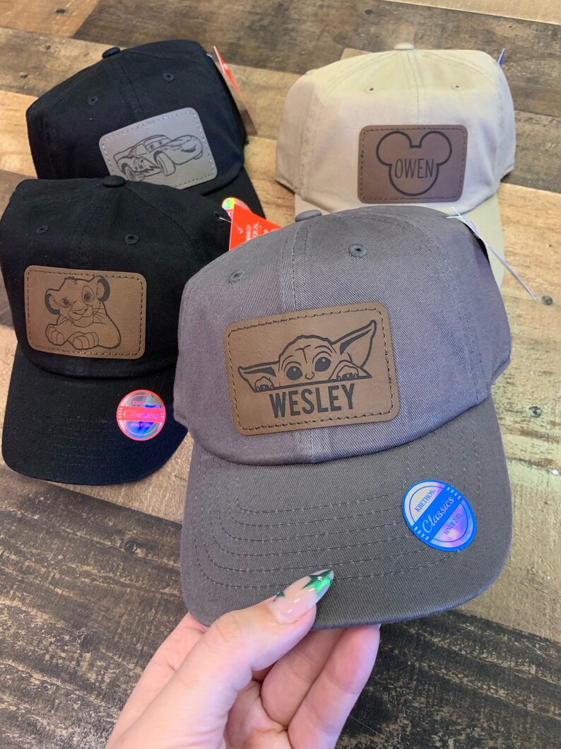 May include: Four baseball caps with personalized patches. The caps are black, beige, gray, and black. The patches feature a cartoon car, Mickey Mouse ears, a baby Yoda, and a lion. The patches are all brown leather with white stitching. The caps have adjustable straps.