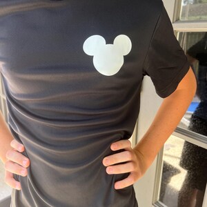 May include: Black short-sleeved shirt with a white Mickey Mouse head graphic on the chest. The shirt has a crew neck and is made of a textured fabric. The person is standing in front of a window.