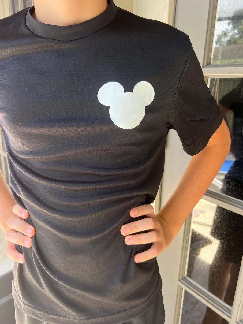 May include: Black short-sleeved shirt with a white Mickey Mouse head graphic on the chest. The shirt has a crew neck and is made of a textured fabric. The person is standing in front of a window.