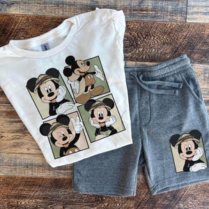 May include: A white t-shirt with a multi-panel design featuring Mickey Mouse in a safari outfit. The shirt is paired with gray sweatpants with a single Mickey Mouse graphic on the left leg.