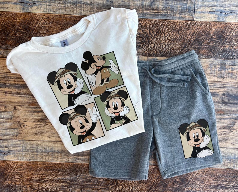 May include: A white t-shirt with a multi-panel design featuring Mickey Mouse in a safari outfit. The shirt is paired with gray sweatpants with a single Mickey Mouse graphic on the left leg.