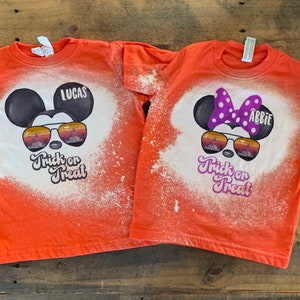 Puede incluir: Dos camisetas naranjas con dise&ntilde;os de lej&iacute;a de Mickey Mouse y Minnie Mouse con gafas de sol. Las camisetas dicen "Trick or Treat" y tienen los nombres "Lucas" y "Abbie" en ellas.