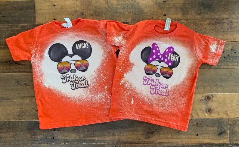 Puede incluir: Dos camisetas naranjas con dise&ntilde;os de lej&iacute;a de Mickey Mouse y Minnie Mouse con gafas de sol. Las camisetas dicen "Trick or Treat" y tienen los nombres "Lucas" y "Abbie" en ellas.