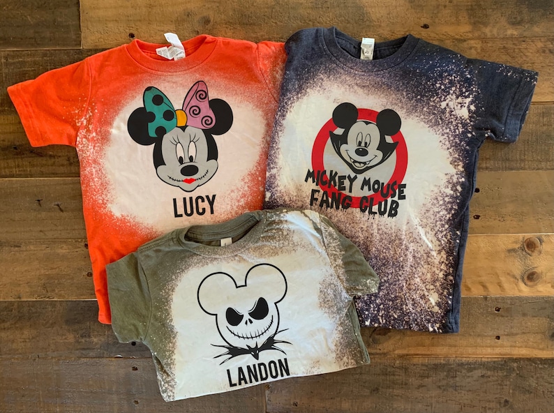 Puede incluir: Tres camisetas blanqueadas con dise&ntilde;os de tem&aacute;tica de Halloween. La camiseta naranja presenta un dise&ntilde;o de Mickey Mouse con el texto "Lucy". La camiseta azul presenta un dise&ntilde;o de Mickey Mouse con el texto "Mickey Mouse Fang Club". La camiseta verde presenta un dise&ntilde;o de Jack Skellington con el texto "Landon".