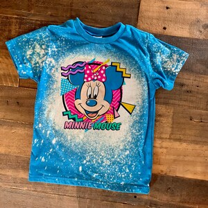 Puede incluir: Una camiseta azul con un dise&ntilde;o salpicado de lej&iacute;a que presenta una imagen de dibujos animados de Minnie Mouse con el texto "Minnie Mouse" debajo.