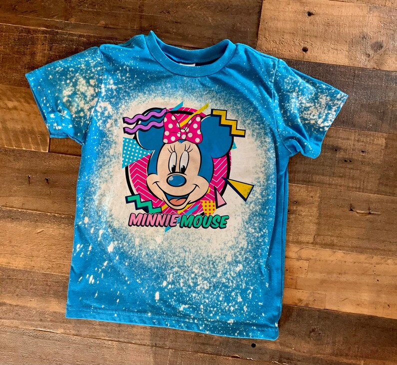 Puede incluir: Una camiseta azul con un dise&ntilde;o salpicado de lej&iacute;a que presenta una imagen de dibujos animados de Minnie Mouse con el texto "Minnie Mouse" debajo.