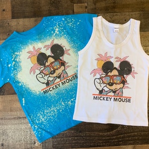 Puede incluir: Dos camisetas con una imagen de dibujos animados de Mickey Mouse con gafas de sol y una camisa hawaiana. Las camisetas tienen un dise&ntilde;o salpicado de lej&iacute;a y el texto "Mickey Mouse" est&aacute; impreso en ambas camisetas.