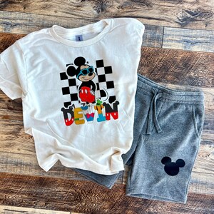 May include: White t-shirt with a black and white checkered background, a cartoon Mickey Mouse wearing sunglasses, and the name "DEVIN" in colorful letters. Gray sweatpants with a black Mickey Mouse silhouette.
