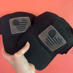 May include: Two black baseball caps with a leather patch featuring a stylized American flag design. The flag design incorporates a Mickey Mouse head shape with stars and stripes. The caps are held against a coral-coloured background.