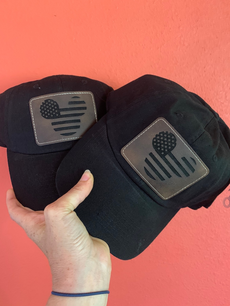 May include: Two black baseball caps with a leather patch featuring a stylized American flag design. The flag design incorporates a Mickey Mouse head shape with stars and stripes. The caps are held against a coral-coloured background.