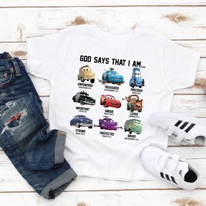 May include: White t-shirt with a Cars themed design. The shirt features different Cars characters with bible verses and the text "God says that I am..."