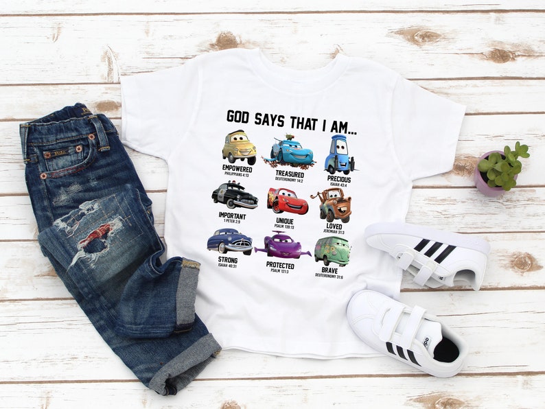 May include: White t-shirt with a Cars themed design. The shirt features different Cars characters with bible verses and the text "God says that I am..."