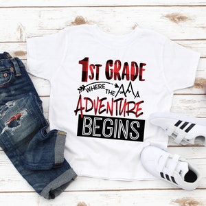 May include: White t-shirt with red and black text that says "1st Grade Where the Adventure Begins". The text is in a buffalo plaid pattern. The shirt is laid out on a white wooden surface with a pair of white sneakers and blue jeans.