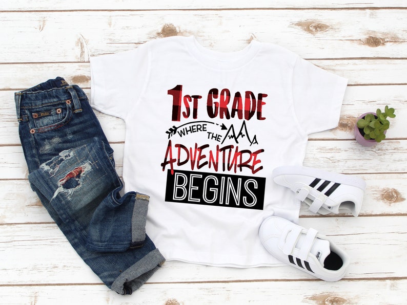 May include: White t-shirt with red and black text that says "1st Grade Where the Adventure Begins". The text is in a buffalo plaid pattern. The shirt is laid out on a white wooden surface with a pair of white sneakers and blue jeans.