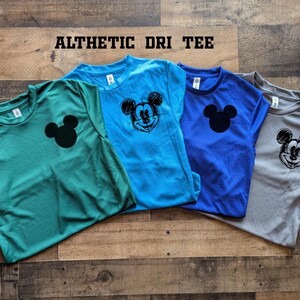 May include: Four athletic dri-fit tees in teal, light blue, royal blue, and gray. Each shirt features a black Mickey Mouse graphic. The shirts are displayed on a wooden surface. The text "ALTHETIC DRI TEE" is at the top.